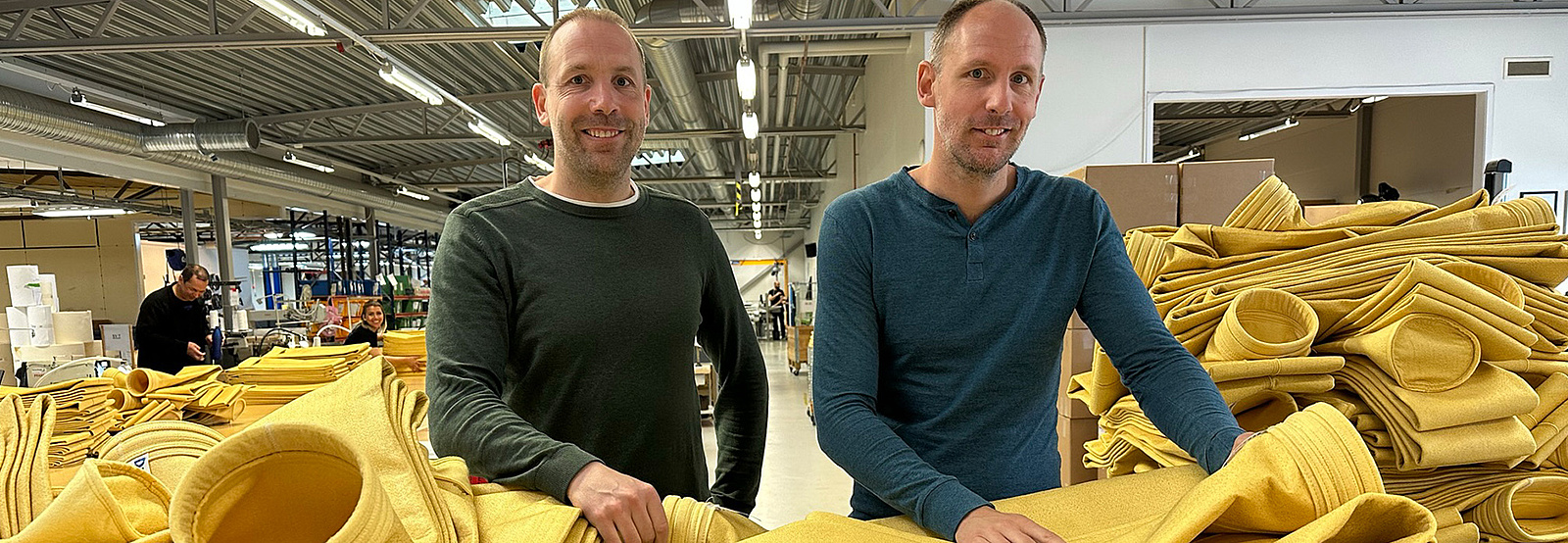 Två leende män står i en fabrikslokal, omringade av gula filterpåsar. 