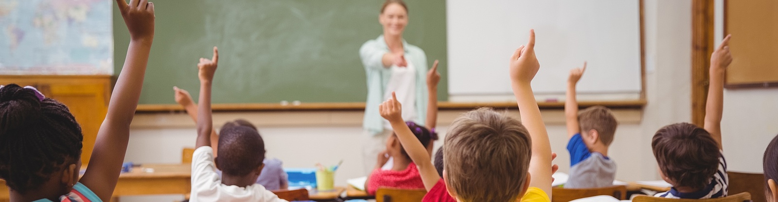 En kvinnlig lärare står leendes framför en grön tavla och pekar mot några barn som räcker upp handen. 