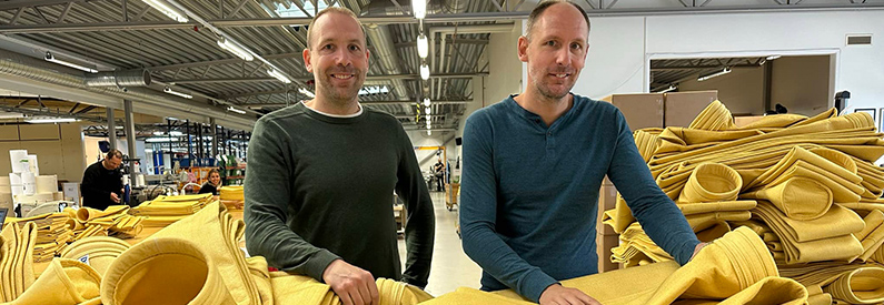 Två leende män står i en fabrikslokal, omringade av gula filterpåsar.